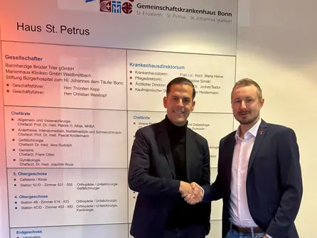 Zwei Männer in Anzügen schütteln sich vor einer Wand mit Informationsplakaten die Hand