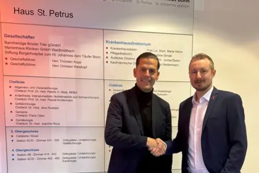 Zwei Männer in Anzügen schütteln sich vor einer Wand mit Informationsplakaten die Hand