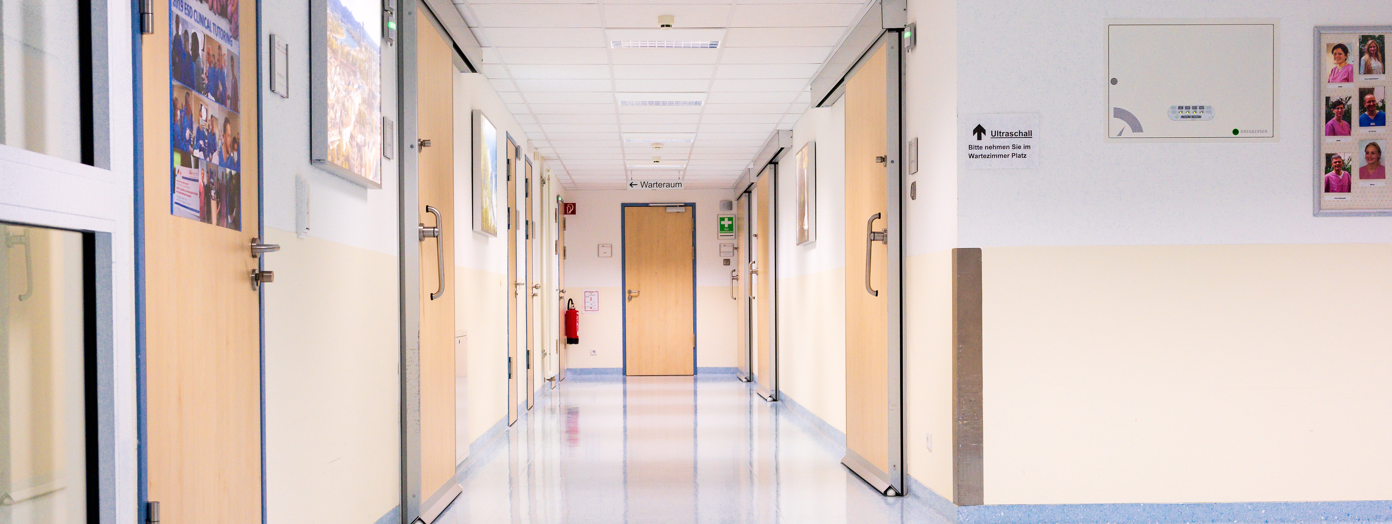 Heller Krankenhausflur mit mehreren geschlossenen Holztüren, weißen Wänden und blauem Linoleumboden, Notausgangsschild an der Decke.