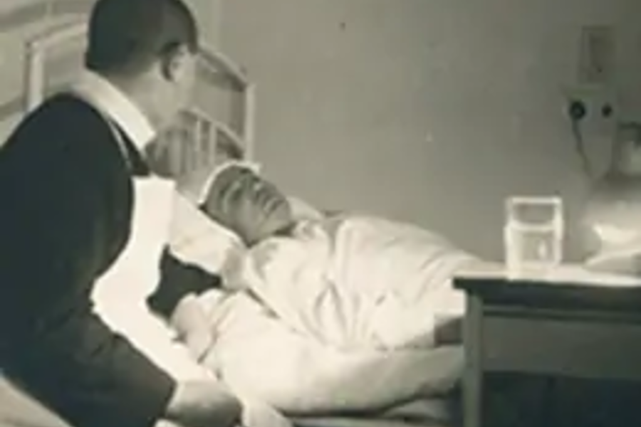 Krankenpfleger mit Schürze beugt sich über im Bett liegenden Patienten in einem historischen Krankenhauszimmer