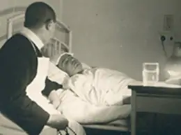 Krankenpfleger mit Schürze beugt sich über im Bett liegenden Patienten in einem historischen Krankenhauszimmer