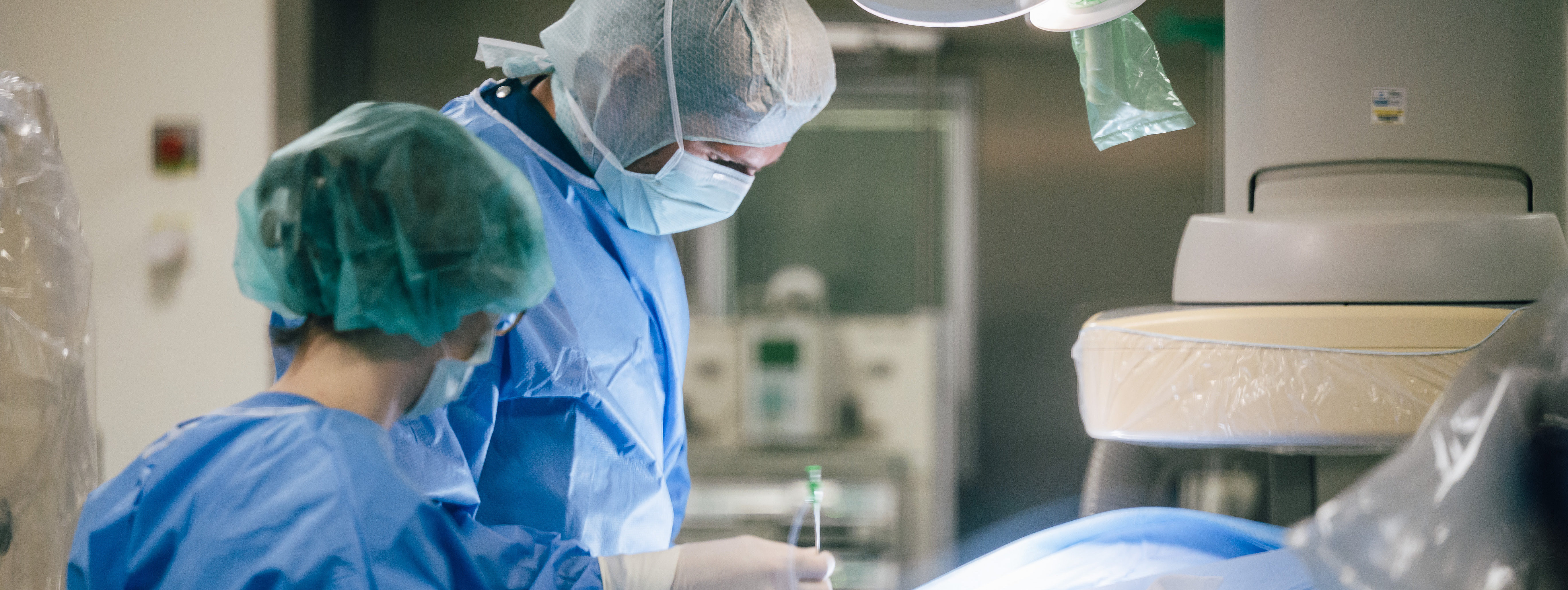 Zwei medizinische Fachkräfte in OP-Kleidung führen einen Eingriff an einem Patienten durch, medizinische Geräte im Hintergrund sichtbar.