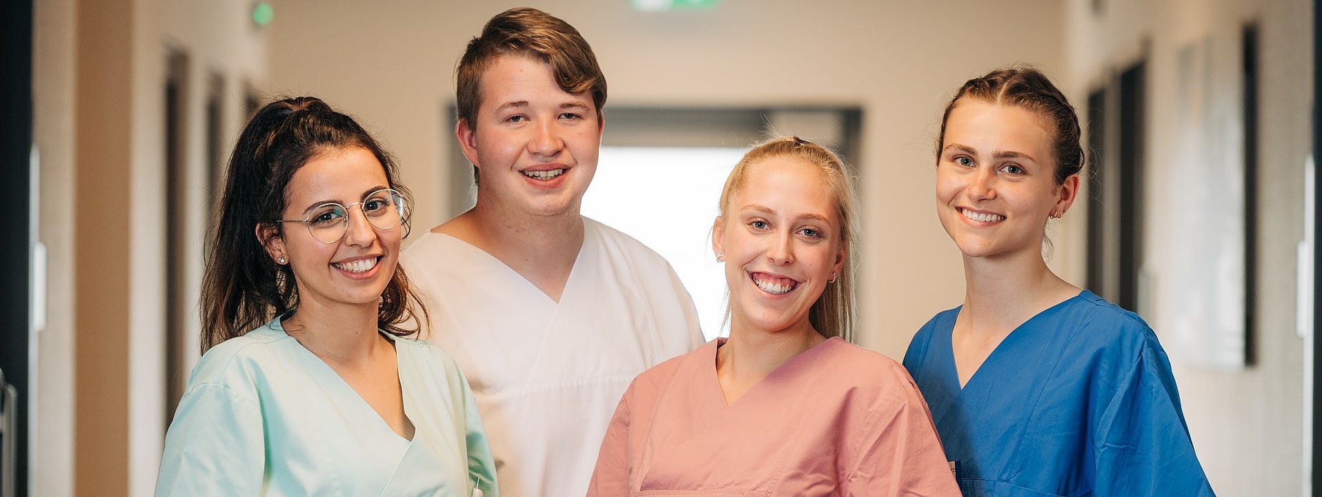 Vier Personen in Krankenhausflur tragen medizinische Kittel in verschiedenen Farben und haben die Hände in den Taschen