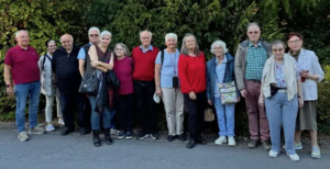 Gruppe älterer Menschen steht auf einem Gehweg vor dichtem Grün