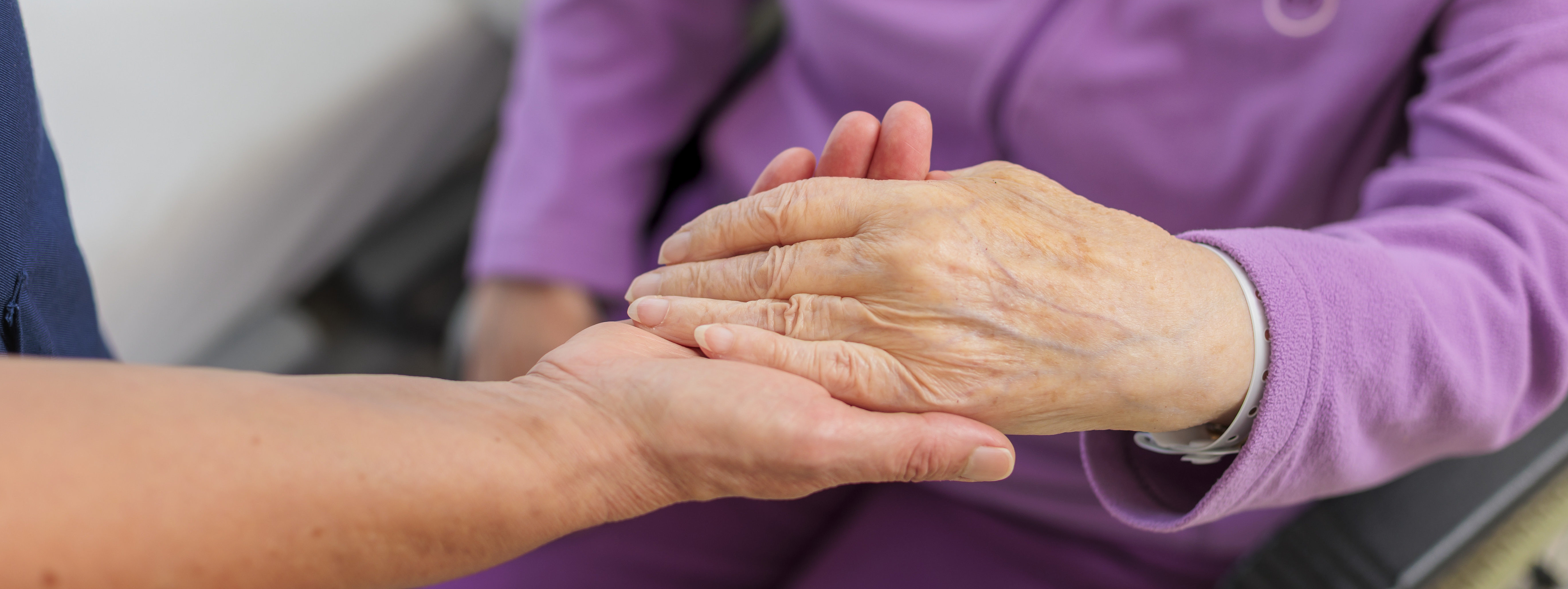 Zwei Hände, eine ältere Hand liegt auf einer jüngeren Hand, ältere Person trägt lila Kleidung und sitzt im Rollstuhl.