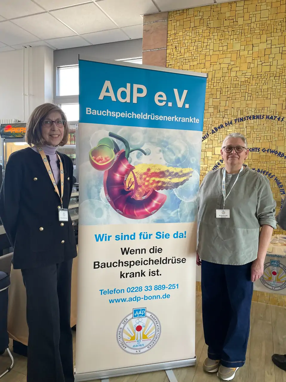 Roll-up-Banner von AdP e.V. mit Illustration einer Bauchspeicheldrüse und Text zu Bauchspeicheldrüsenerkrankungen, zwei Personen stehen daneben