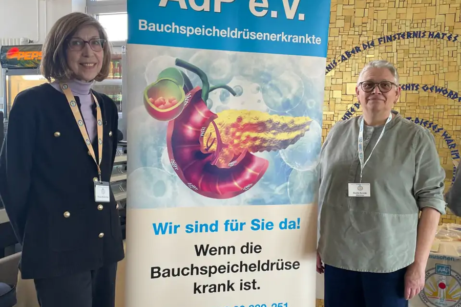 Roll-up-Banner von AdP e.V. mit Illustration einer Bauchspeicheldrüse und Text zu Bauchspeicheldrüsenerkrankungen, zwei Personen stehen daneben
