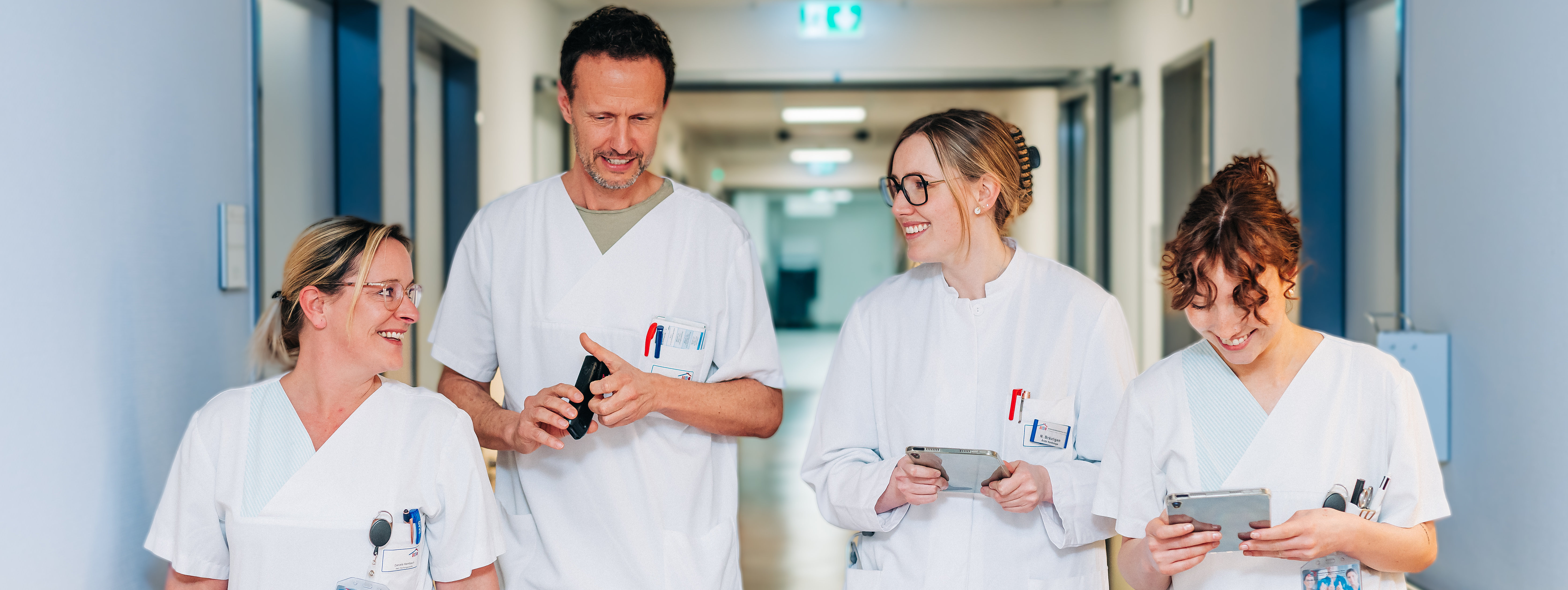 Vier medizinische Fachkräfte in weißen Kitteln und Uniformen gehen einen Krankenhausflur entlang, zwei halten Tablets in der Hand