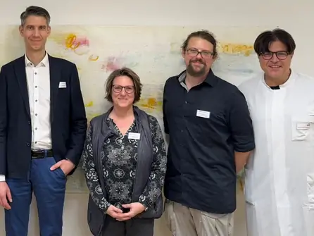 Vier Personen stehen nebeneinander vor einer Wand mit einem großen, farbigen Poster