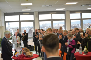 Menschen in einem hellen Raum mit großen Fenstern applaudieren einer Person, die vor einem Tisch mit Geschenken steht.