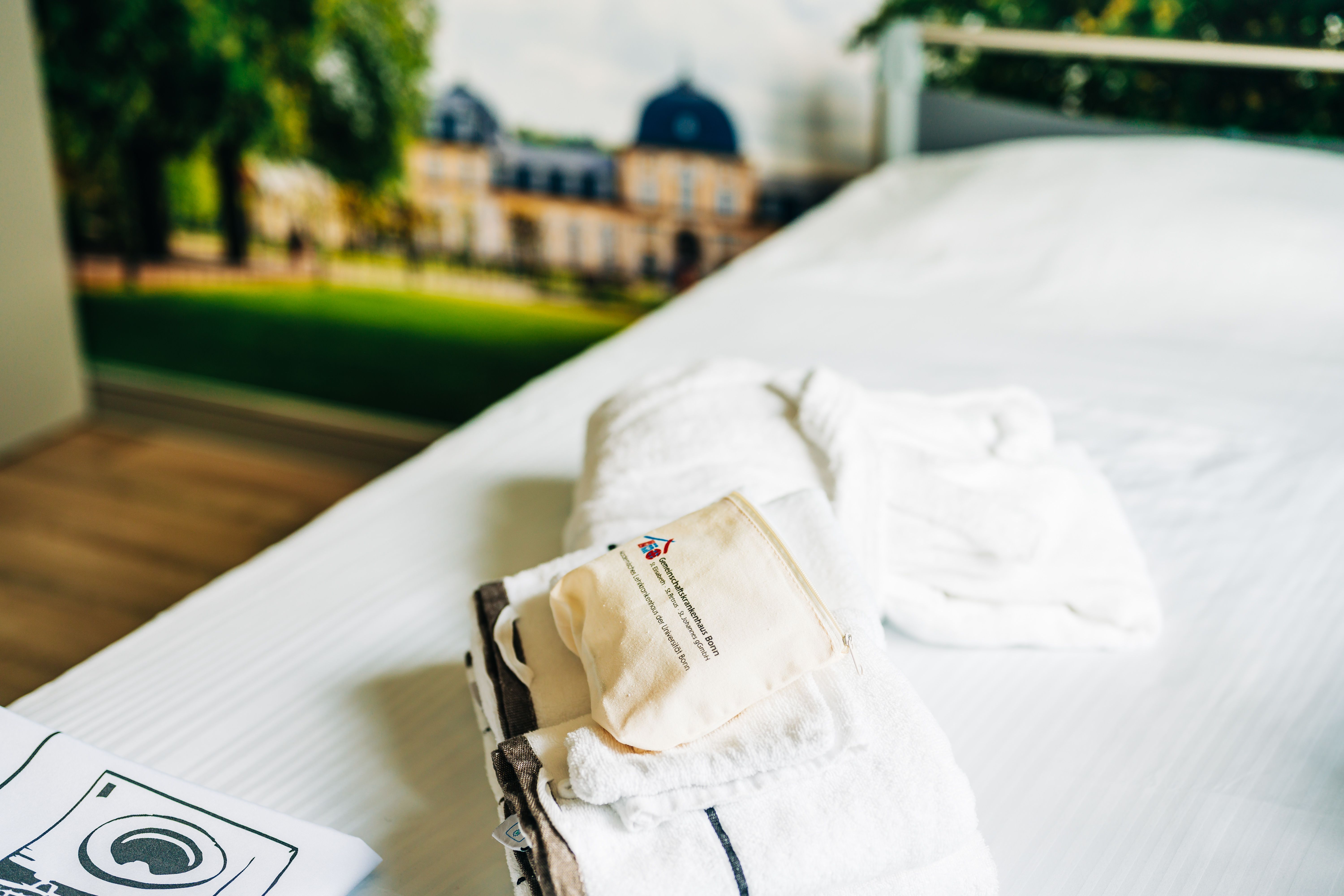 Gefaltete weiße Handtücher und ein Beutel auf einem weißen Bett in einem Hotelzimmer mit Blick auf ein Schloss im Hintergrund
