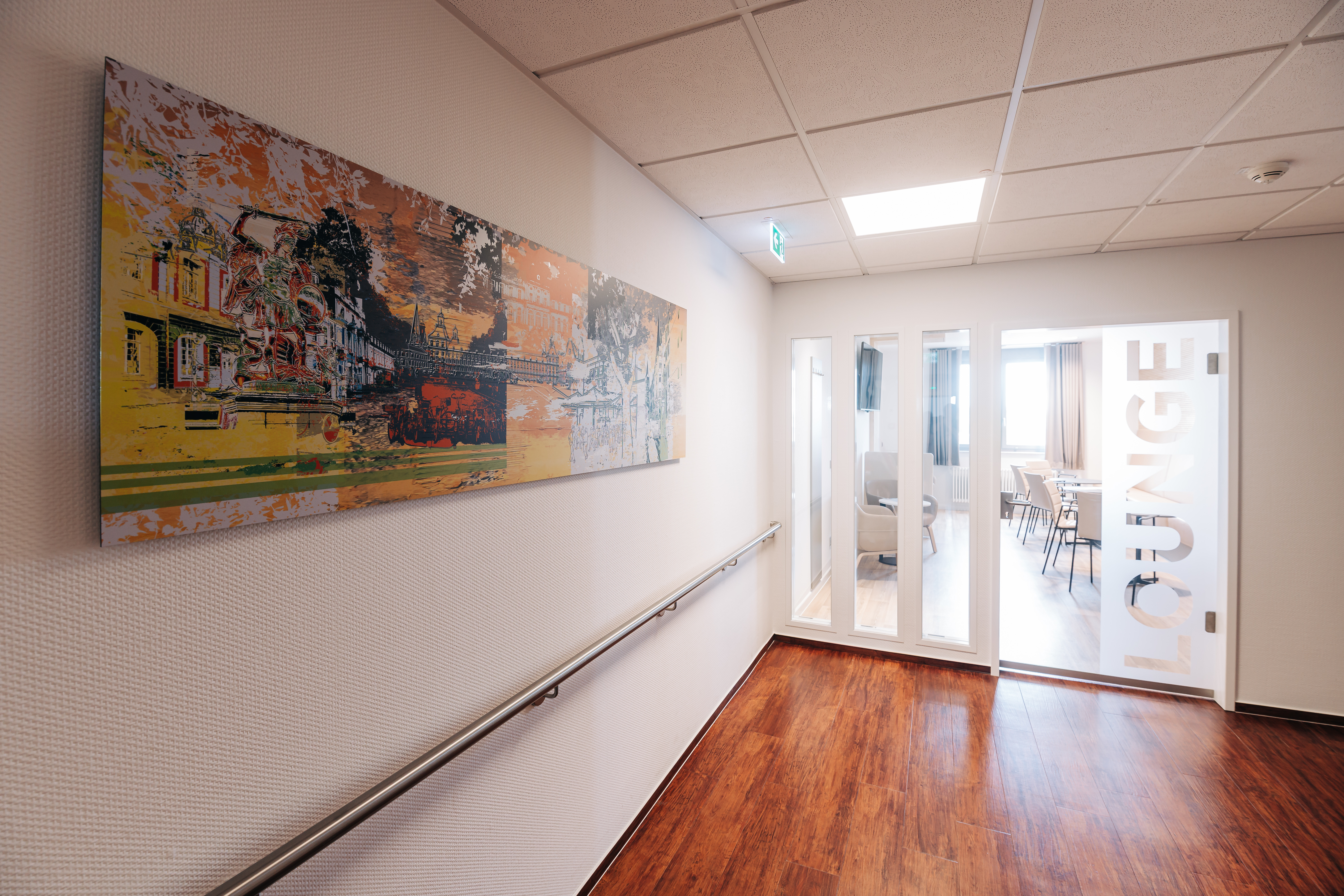 Flur mit Handlauf und abstraktem Wandbild, am Ende Glastür mit Schriftzug 'LOUNGE' zu einem Raum mit Stühlen und Tischen
