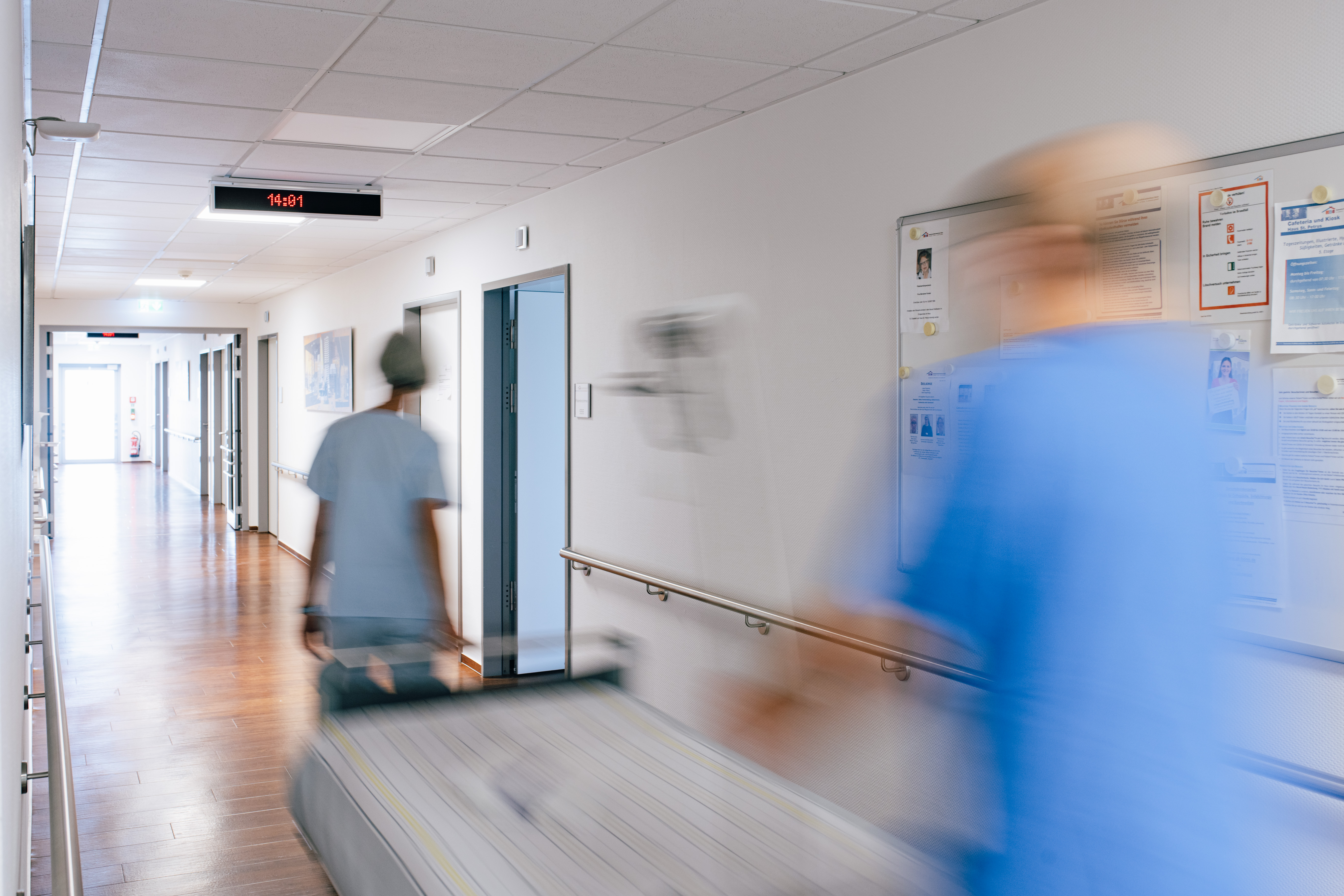 Unscharfe Aufnahme eines Krankenhausflurs mit zwei Personen in medizinischer Kleidung, eine schiebt ein Bett.