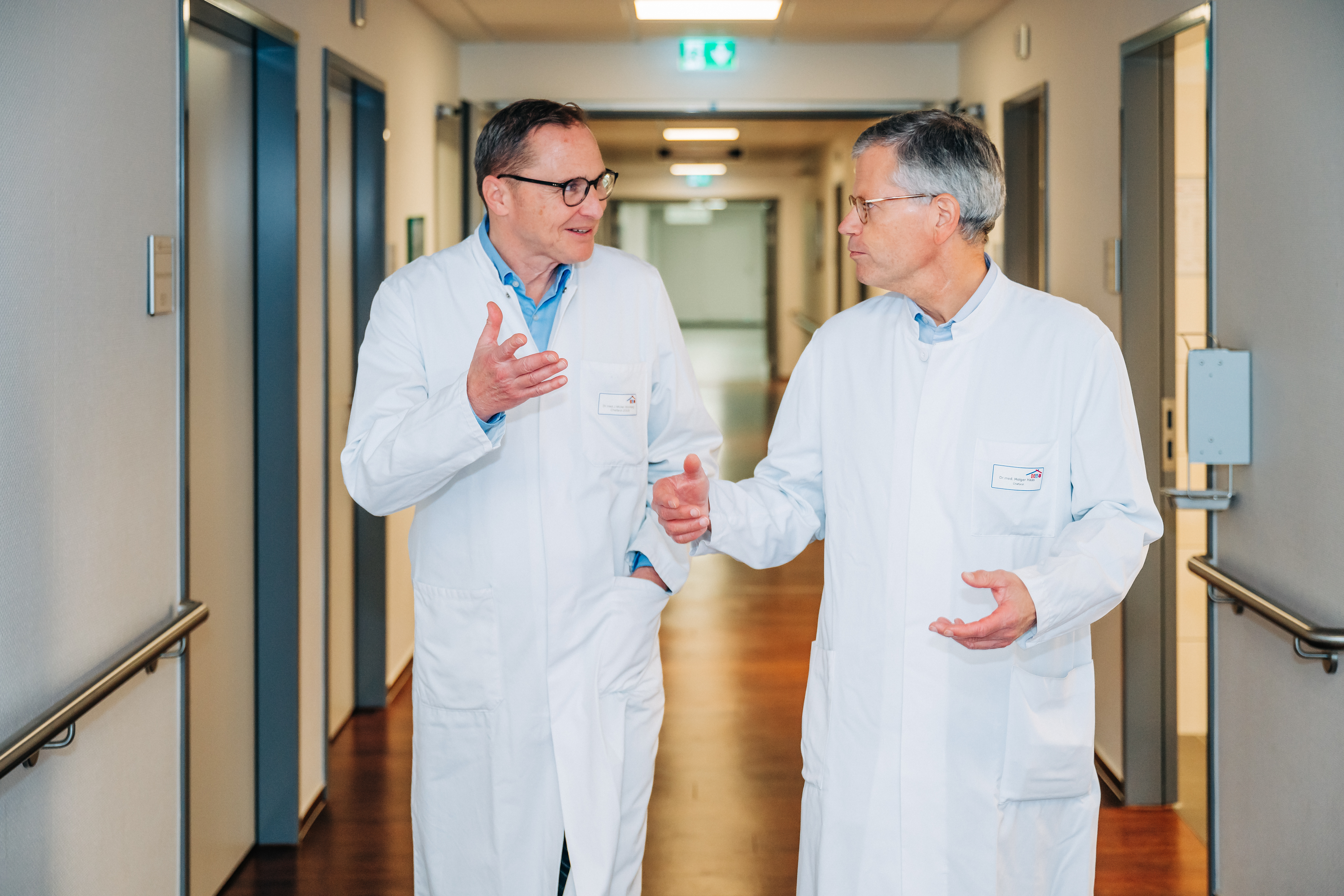 Zwei Männer in weißen Arztkitteln gehen und unterhalten sich in einem Krankenhausflur.