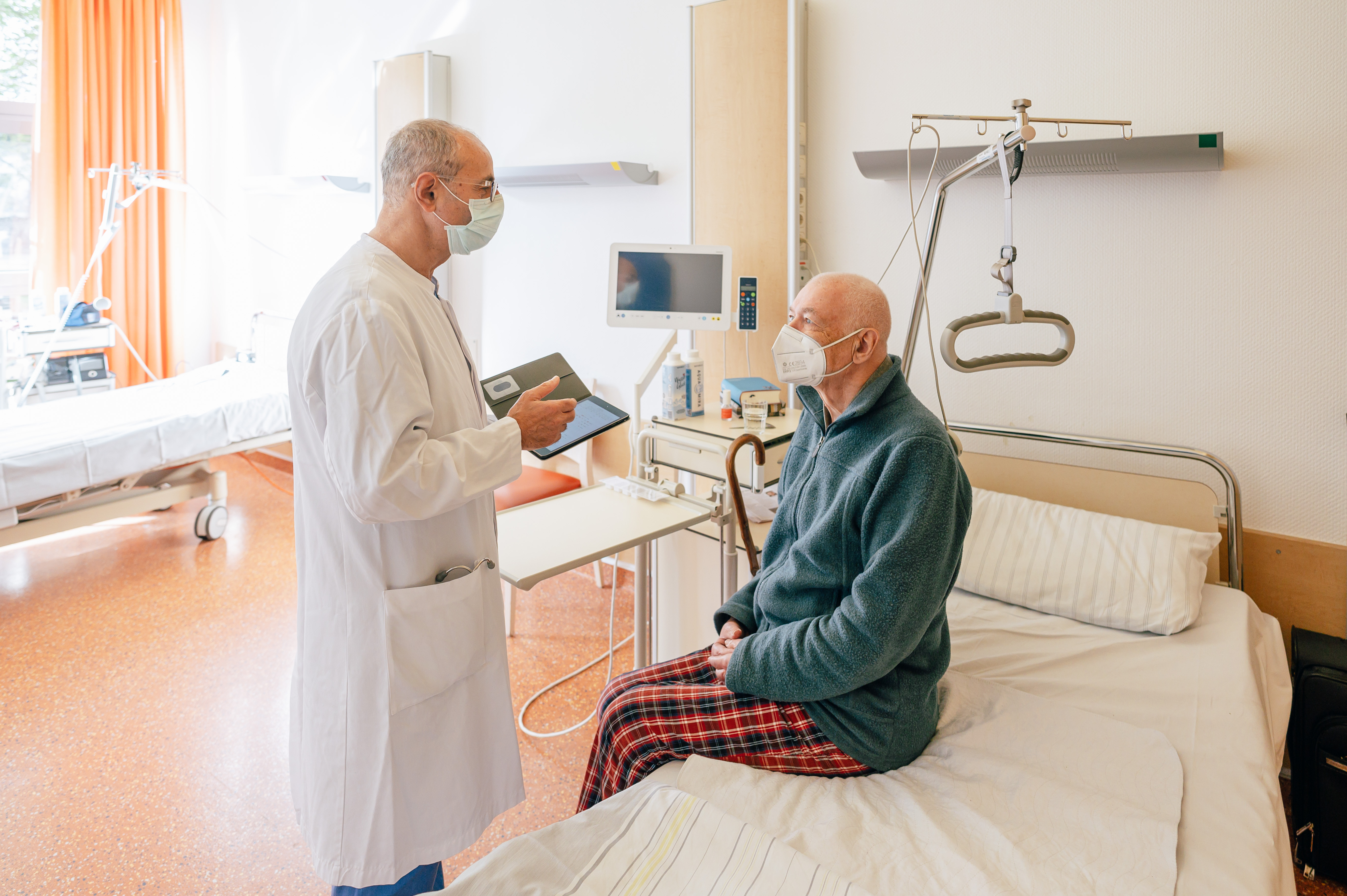Ein Arzt im weißen Kittel steht neben einem Patienten, der auf einem Krankenhausbett sitzt, in einem hellen Patientenzimmer.