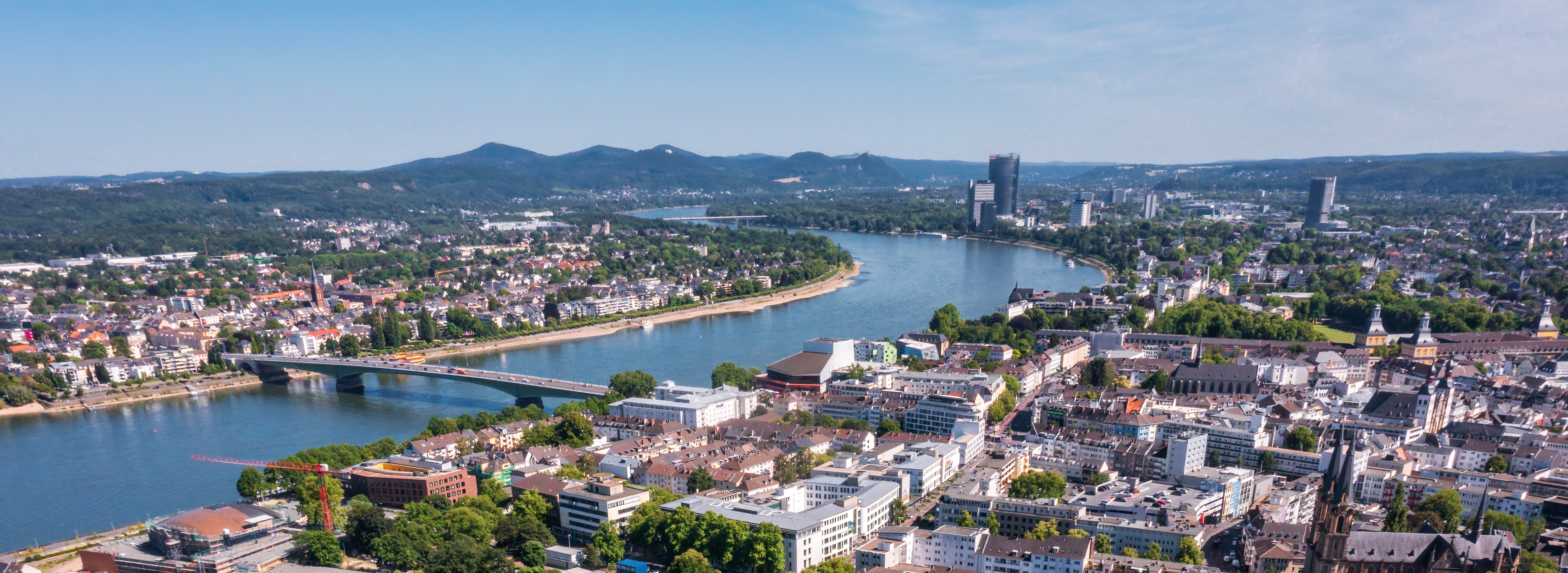 Luftaufnahme einer Stadt mit Fluss, Brücke und mehreren Gebäuden vor bewaldeten Hügeln im Hintergrund